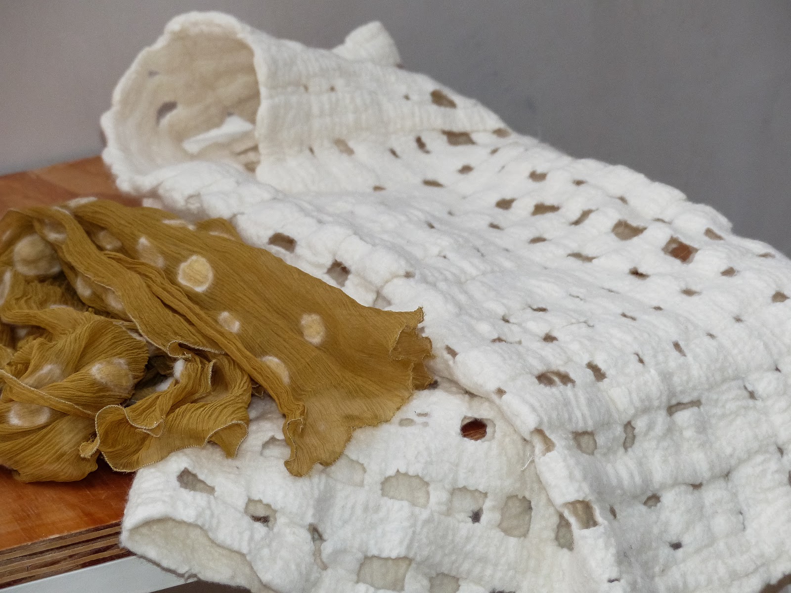 Dos prendas de nuno fieltro sobre una mesa de madera: una blanca calada en retícula y una bufanda de seda mostaza con lunares claros.